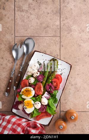 Asparagi, pomodoro, lattuga, mozzarella, sesamo nero, lino, insalata di olio d'oliva e uovo bollito morbido su piastra rettangolare in ceramica su tavola di piastrelle grigio chiaro Foto Stock