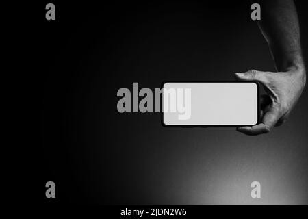 Immagine in bianco e nero della mano dell'uomo che tiene lo smartphone nero orizzontalmente con schermo bianco vuoto isolato su sfondo scuro con illuminazione drammatica A. Foto Stock