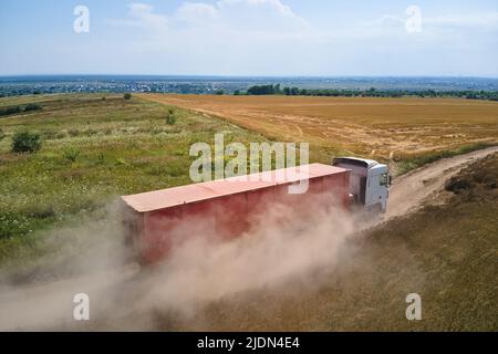 Vista aerea del cargo truck che guida su strada sterrata tra campi di grano agricolo facendo molta polvere. Trasporto di grano dopo essere stato raccolto da Foto Stock