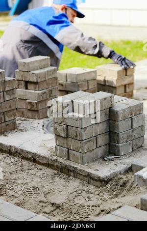 Il muratore in abiti da lavoro si siede sul marciapiede e stende le lastre di pavimentazione. Vista dell'uomo in attività all'aria aperta. Costruttore professionista fa organizzazione del territorio giorno d'estate. Scena reale. Foto Stock