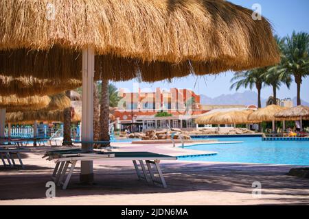 Sedie a sdraio vuote sotto ombrelloni di paglia sul lato piscina nel resort tropicale. Vacanza estiva e concetto di fuga Foto Stock