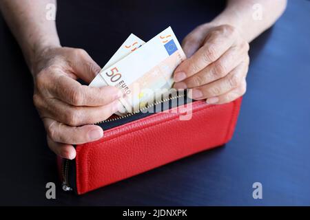 Donna anziana prende le banconote in euro dal portafoglio rosso, rughe donna mani primo piano. Concetto di previdenza e assistenza, risparmio, pensionamento Foto Stock