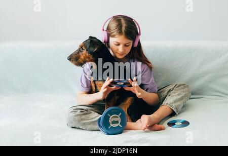 Ragazza felice adolescente in t-shirt lilla in cuffie rosa ascolta la musica e ha divertimento a giocare con il cane Foto Stock