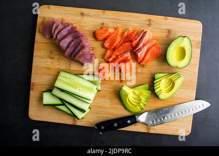 Affettare pesce fresco e verdure per fare sushi: Pesce affettato, avocado e cetriolo su un tagliere di bambù Foto Stock