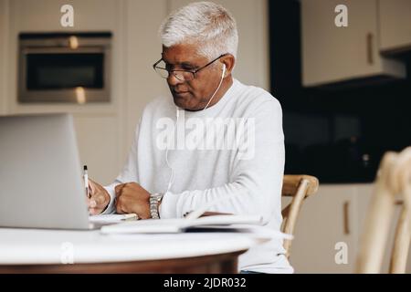 Uomo d'affari anziano che scrive i suoi piani in un taccuino mentre si siede nel suo ufficio domestico. Uomo d'affari maturo che usa un laptop mentre lavora da casa. Foto Stock