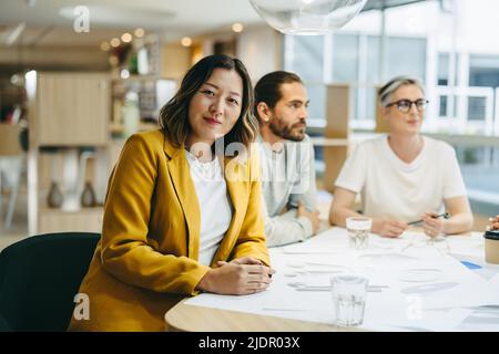 Donna d'affari etnica che guarda la macchina fotografica mentre si siede in una riunione con i suoi colleghi. Gruppo di professionisti del design innovativi che brainstorming whil Foto Stock