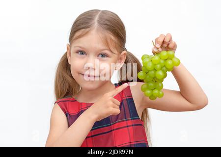 Bella ragazza caucasica piccola di 6 anni che tiene mazzo di uve verdi e punto su di esso da indice dito su sfondo bianco guardando la macchina fotografica Foto Stock