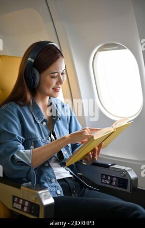 L'attraente passeggero asiatico millenario si rilassa ascoltando la musica sulle moderne cuffie senza fili e leggendo un libro durante un volo. Viaggio, tour Foto Stock