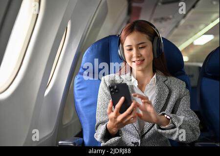 Una donna d'affari asiatica di successo e attraente che utilizza lo smartphone, ascolta la musica con le sue cuffie wireless mentre viaggia in aereo. Foto Stock