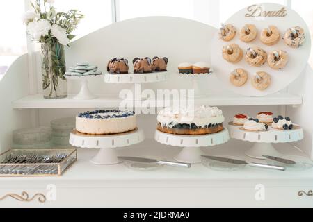 Serve ciambelle decorate, torte alla bacca, biscotti e cupcake cremosi su vassoio, sfondo bianco. Dolci al forno, festa dell'evento. Pasticceria fatta in casa Foto Stock