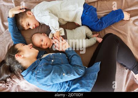 La mamma felice coccola con il bambino e il fratello maggiore su una coperta nel soggiorno Foto Stock