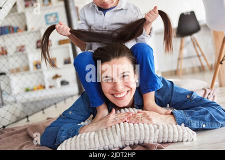Felice madre sdraiata sul pavimento e lasciando che suo figlio le tiri i capelli Foto Stock