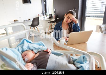 Madre esausta al PC portatile in ufficio con il bambino che dorme sul letto in primo piano Foto Stock