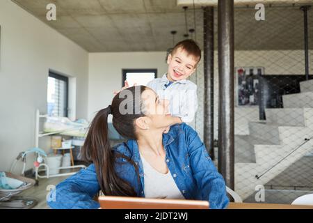 Il bambino felice vuole giocare e disturba la madre sul computer in casa d'ufficio in soggiorno Foto Stock