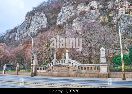 Il ripido pendio della collina di Gellert con un piccolo ponte in pietra intagliato della Cappella della roccia ai suoi piedi, Budapest, Ungheria Foto Stock