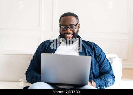 Uomo afroamericano sorridente in camicia casual sopra t-shirt e jeans seduti sul divano in appartamento moderno, ragazzo nero freelancer, imprenditore, maschio studente utilizzando il laptop, godendo di chat online Foto Stock