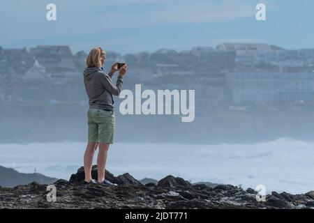 Una villeggiatrice femminile in piedi sulle rocce sulla costa utilizzando il suo smartphone per fotografare Fistral Bay a Newquay in Cornovaglia nel Regno Unito. Foto Stock