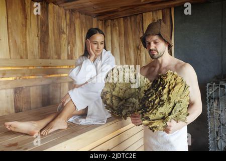 Uomo muscoloso con ginestra nella sauna e la sua donna seduta sullo sfondo. Bella coppia nella sauna Foto Stock