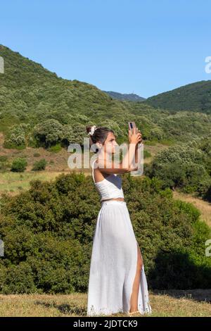 Vista verticale di una giovane donna turistica in abito bianco e gonna lunga scattando una foto con il suo smartphone durante le vacanze estive Foto Stock
