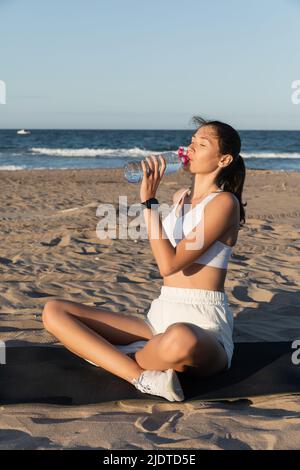 giovane donna che tiene la bottiglia con acqua mentre si siede sul tappeto fitness vicino al mare Foto Stock