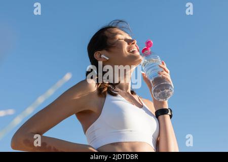 vista ad angolo basso di donna allegra in auricolare wireless che tiene la bottiglia con acqua contro il cielo blu Foto Stock