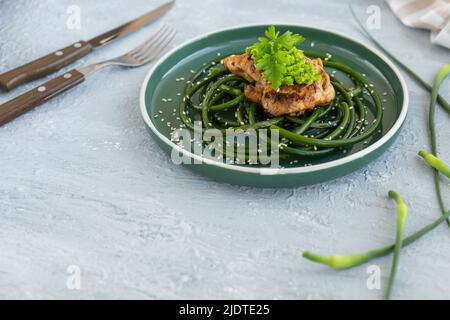 Bistecca di pollo fritta con frecce all'aglio e semi di sesamo in piatto verde su tavola grigia in cemento Foto Stock