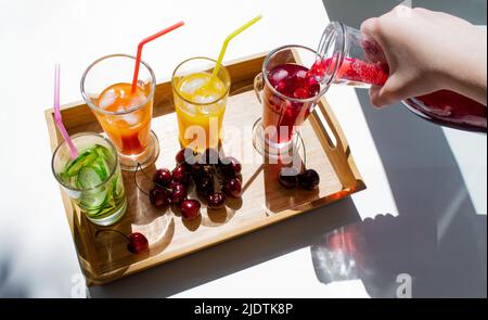 Vassoio di legno con bevande colorate, ciliegie. Decanter con i resti di succo rosso. Finestra, estate. Foto Stock