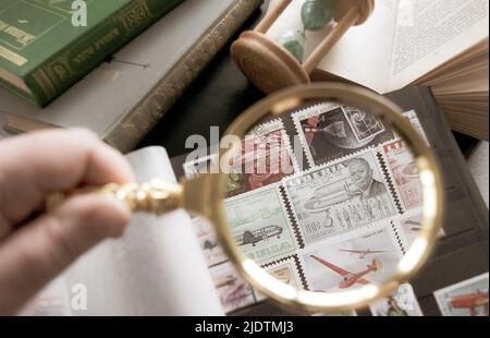 Mosca, Russia, Marzo 2022: Hobby: Raccolta di francobolli. Guardando il francobollo da 50 cent della Liberia attraverso una lente d'ingrandimento dorata. Vecchi libri e una clessidra Foto Stock