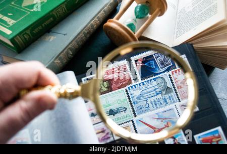 Mosca, Russia, Marzo 2022: Hobby: Raccolta di francobolli. Guardando il francobollo da 50 cent della Liberia attraverso una lente d'ingrandimento dorata. Vecchi libri e una clessidra Foto Stock