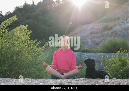 Giovane donna in maglione rosa seduta in posizione di loto e meditating nella natura bella all'alba con il suo cane nero di recupero di labrador che giace accanto Foto Stock