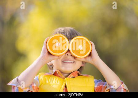 Sorprendo il bambino che tiene le fette di frutta arancione come gli occhiali da sole. Ragazzo divertente contro autunno sfocato sfondo. Felice ragazzo in autunno all'aperto. Eati sani Foto Stock