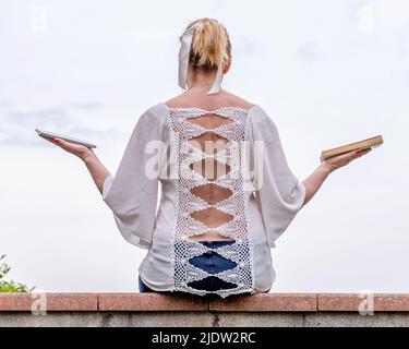 Una donna seduta su un muro tiene una tavoletta in una mano e un libro nell'altra Foto Stock