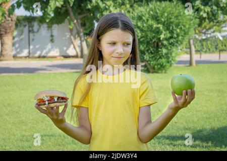 Una ragazza adolescente sceglie tra un hamburger e una mela all'aperto nel parco. Foto Stock