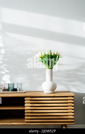 Interno casa scandinavo con bouquet primavera di fiori tulipano bianco in vaso di ceramica, vassoio con candele in piedi su un mobile di legno. Desi minimalisti Foto Stock