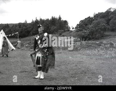 1950s, storico, un piper scozzese in abito regimentale completo in piedi con le sue cornamuse, in alto terreno nelle Highlands, Scozia, Regno Unito. Foto Stock