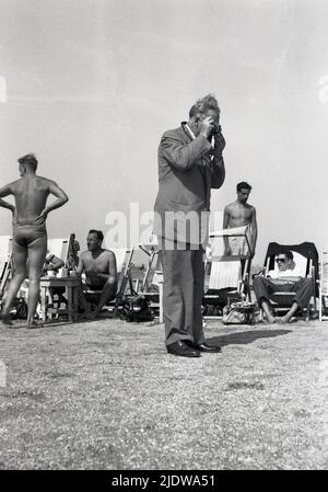 1950s, storico, un gentleman in un vestito e cravatta in piedi su una spiaggia facendo una fotografia, Napoli, Italia Foto Stock