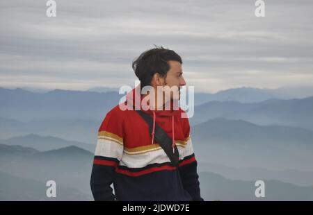 Vita in su di un ragazzo indiano bello osservare giovane guardando lateralmente mentre in piedi sullo sfondo delle montagne Foto Stock