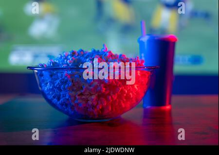 Una partita di calcio su un grande schermo TV, una ciotola di popcorn e una bevanda analcolica in un vetro di plastica su un tavolo di legno. Background sportivo. Guardare lo sport pro Foto Stock