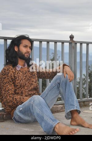Un affascinante ragazzo indiano giovane dai capelli lunghi e bearded che guarda lateralmente, seduto con appoggiarsi sulla barriera di sicurezza sullo sfondo delle montagne Foto Stock