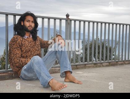 Un giovane indiano dall'aspetto piacevole con capelli lunghi e barba che guarda la macchina fotografica mentre si siede con appoggiarsi sulla barriera di sicurezza sullo sfondo delle montagne Foto Stock