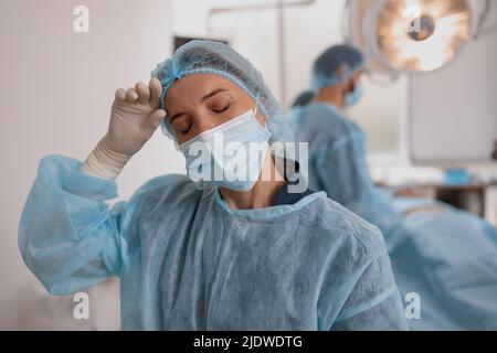 Primo piano di un chirurgo stanco in maschera in piedi in sala operatoria dopo un intervento chirurgico importante Foto Stock