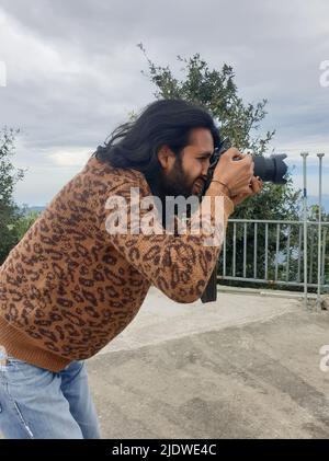 Vista laterale di un giovane indiano con capelli lunghi e bearded che scatta foto con la macchina fotografica mentre si alza all'esterno Foto Stock