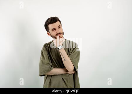 Bell'uomo caucasico che indossa la t-shirt kaki posa isolata su sfondo bianco pensando e toccando il mento mentre si guarda da parte. Soluzione del problema Foto Stock