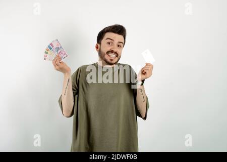 Uomo casual felice che indossa t-shirt posa isolato su sfondo bianco tenendo carta di credito e soldi lira turco, viso sorridente e guadagni felici Foto Stock