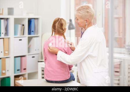 Vista posteriore di pediatra esperto maturo in camice da laboratorio utilizzando stetoscopio durante l'esame del bambino su appuntamento in ufficio Foto Stock