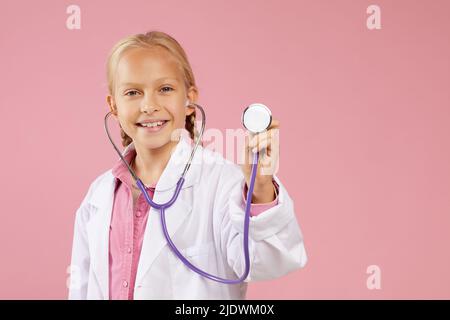 Ritratto di ragazza piccola felice in camice da laboratorio in piedi su sfondo rosa e utilizzando stetoscopio mentre sogna di diventare medico Foto Stock