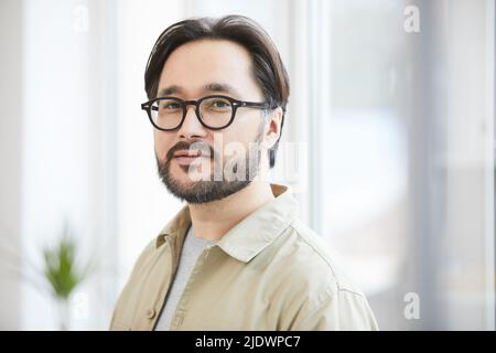 Ritratto di contenuto fiducioso hipster giovane asiatico ragazzo con la barba indossare camicia e occhiali in ufficio moderno Foto Stock