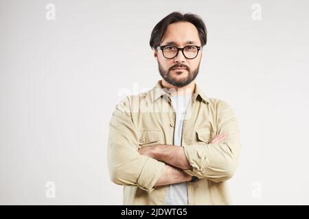 Ritratto del giovane asiatico serio e bello in maglia khaki con occhiali eleganti che incrociano le braccia su sfondo grigio Foto Stock