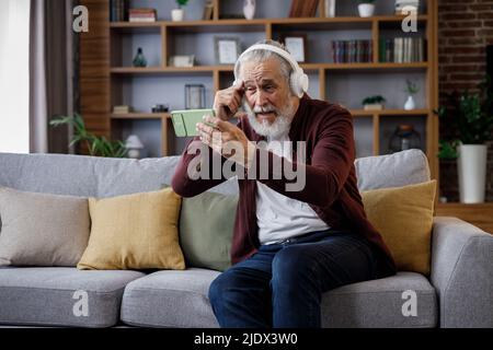 Felice uomo anziano dai capelli grigi in cuffie bianche che tengono lo smartphone e ridono. Vecchio uomo seduto sul divano a casa guardando contenuti divertenti Foto Stock