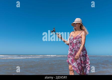 Donna bionda in cappello e abito estivo che fa una foto mentre passeggi sulla spiaggia Foto Stock